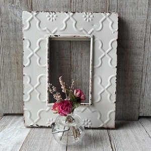 May include: A distressed white metal picture frame with a repeating floral and geometric pattern. The frame surrounds an empty rectangular space. A small glass vase with dried flowers sits in front of the frame.
