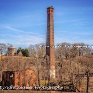 May include: A tall, red brick chimney stands against a blue sky with white clouds. The chimney is surrounded by trees and bushes. The text "Copyright Keastler Photography" appears twice in the image.
