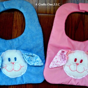 May include: Two baby bibs with bunny faces. One bib is blue with white stars and the other is pink with white polka dots.  A Crafty One, LLC.
