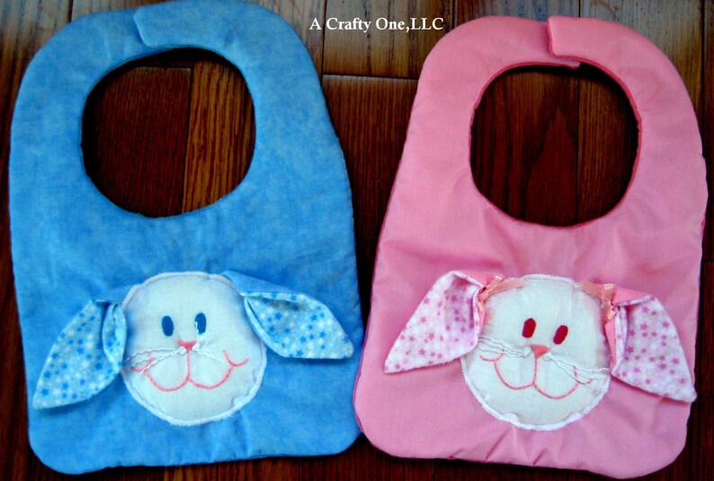 May include: Two baby bibs with bunny faces. One bib is blue with white stars and the other is pink with white polka dots.  A Crafty One, LLC.