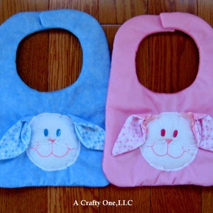 May include: Four baby bibs with bunny faces. The bibs are green, blue, pink, and purple. Each bib has a white bunny face with embroidered eyes, nose, and mouth. The bunny ears are attached to the bib and have a polka dot pattern.