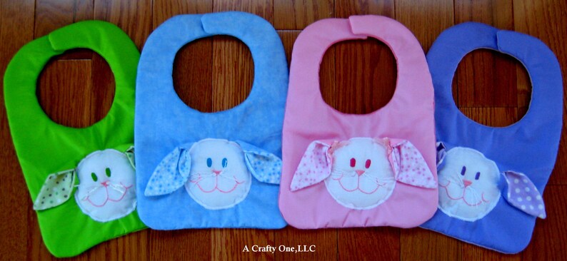 May include: Four baby bibs with bunny faces. The bibs are green, blue, pink, and purple. Each bib has a white bunny face with embroidered eyes, nose, and mouth. The bunny ears are attached to the bib and have a polka dot pattern.
