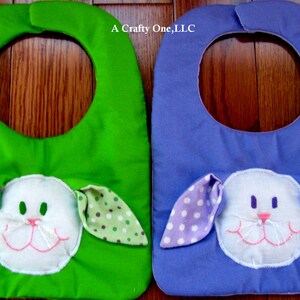 May include: Two baby bibs with a bunny face design. One bib is green with white polka dots and the other is purple with white polka dots. The text "A Crafty One, LLC" is printed on the bibs.