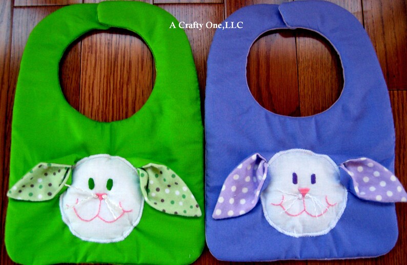 May include: Two baby bibs with a bunny face design. One bib is green with white polka dots and the other is purple with white polka dots. The text "A Crafty One, LLC" is printed on the bibs.