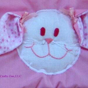 May include: A pink fabric baby blanket with a white felt bunny face appliqu&eacute;. The bunny has pink ears with white stars, pink eyes, and a pink nose.