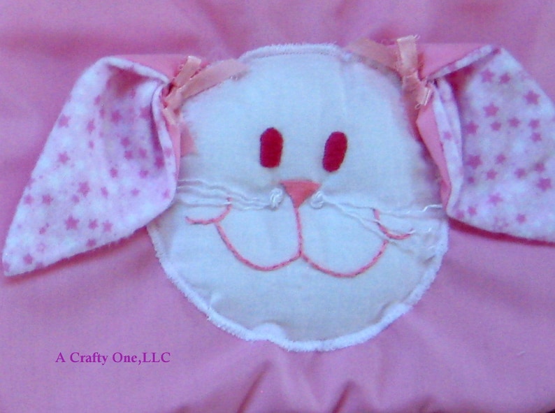 May include: A pink fabric baby blanket with a white felt bunny face appliqu&eacute;. The bunny has pink ears with white stars, pink eyes, and a pink nose.