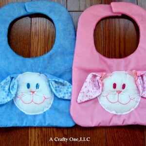 May include: Four baby bibs with bunny faces. The bibs are green, blue, pink, and purple. Each bib has a white bunny face with embroidered details. The text "A Crafty One, LLC" is visible at the bottom of the image.