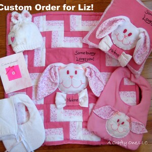 May include: A pink and white baby quilt with a bunny applique and the text "Some bunny loves you!"  A matching pink bib with a bunny applique and the text "Helena". A white hooded towel with pink ties. A pink pillow with a bunny applique and the text "Some bunny loves you!" and "Helena". A pink card with the text "Cute as a button!"