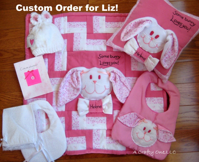 May include: A pink and white baby quilt with a bunny applique and the text "Some bunny loves you!"  A matching pink bib with a bunny applique and the text "Helena". A white hooded towel with pink ties. A pink pillow with a bunny applique and the text "Some bunny loves you!" and "Helena". A pink card with the text "Cute as a button!"