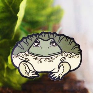 May include: A green and white cartoon frog sticker with a sad expression. The sticker is on a clear surface with green leafy plants in the background.