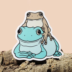 May include: A sticker featuring two frogs, one sitting on top of the other. The bottom frog is blue and the top frog is brown. The frogs are both smiling.