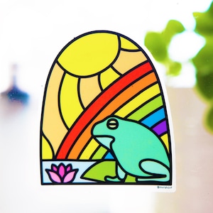 May include: A sticker featuring a green frog sitting on a lily pad with a rainbow and sun behind it. The sticker is in a stained glass style with black outlines.