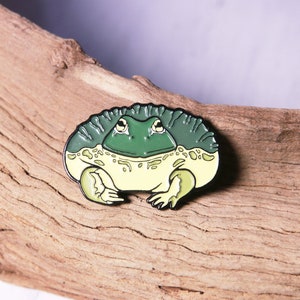 May include: A green and yellow enamel pin of a cartoon frog with a textured skin pattern. The frog is sitting with its legs crossed and has a wide, toothy grin.