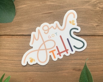 May include: A sticker with the phrase "You Got This" in a playful, handwritten font. The text is in shades of peach, orange, blue, and green, with small floral details. The sticker is set against a wooden backdrop.