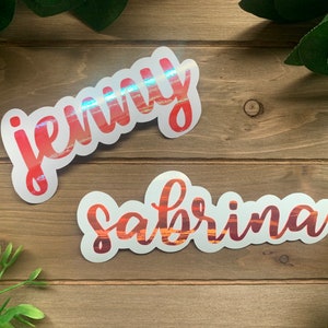 Puede incluir: Dos pegatinas personalizadas con los nombres "Jenny" y "Sabrina" en fuente cursiva roja con un fondo de puesta de sol.
