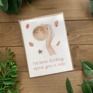 May include: A white card with a watercolor illustration of a hand holding a cup of coffee. The text "I've bean thinking about you a latte" is written in pink on the card.