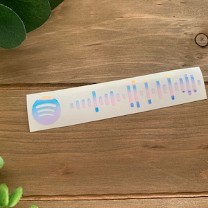May include: A white sticker with a blue and purple gradient Spotify logo and a holographic barcode. The sticker is on a wooden surface.