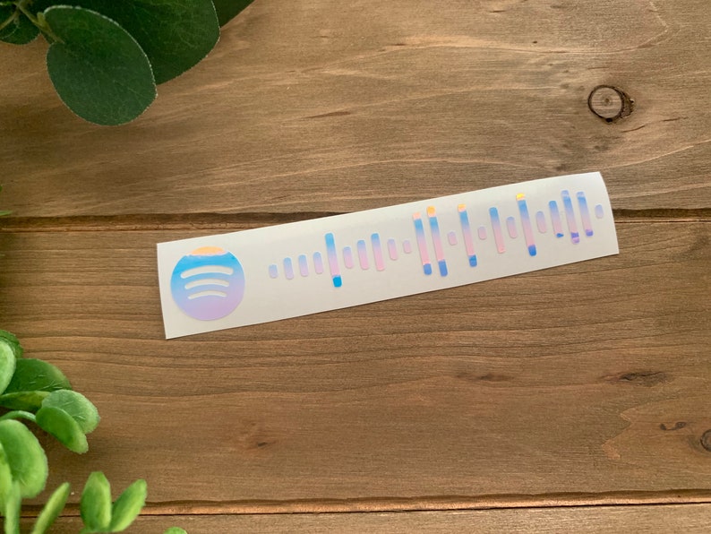 May include: A white sticker with a blue and purple gradient Spotify logo and a holographic barcode. The sticker is on a wooden surface.