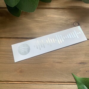 May include: A white sticker with a silver Spotify logo and a silver barcode. The sticker is on a wooden surface.