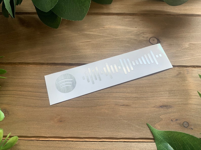 May include: A white sticker with a silver Spotify logo and a silver barcode. The sticker is on a wooden surface.