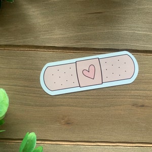 May include: A pink and white band-aid sticker with a small pink heart in the center. The sticker is on a wooden surface.