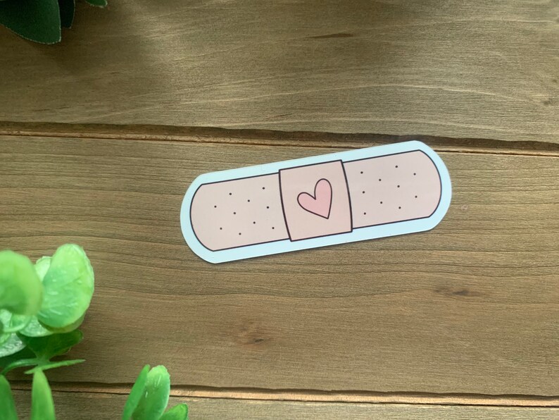 May include: A pink and white band-aid sticker with a small pink heart in the center. The sticker is on a wooden surface.
