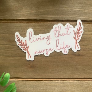 May include: White sticker with pink text that reads "living that nurse life" with two brown leafy sprigs on either side.