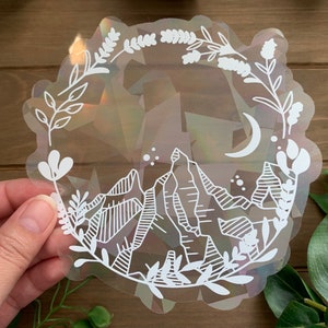 May include: A clear sticker with a white design of a crescent moon, mountains, and floral elements. The sticker has an iridescent background.