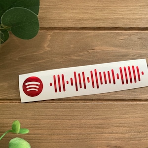 May include: A white sticker with a red Spotify logo and a red barcode. The sticker is on a wooden surface.