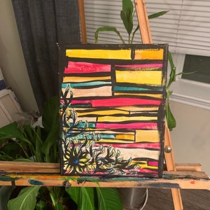 May include: Abstract art painting on canvas. The artwork features a colorful geometric design with yellow, red, and blue horizontal stripes. A black floral design is in the lower left corner. The painting is displayed on a wooden easel.