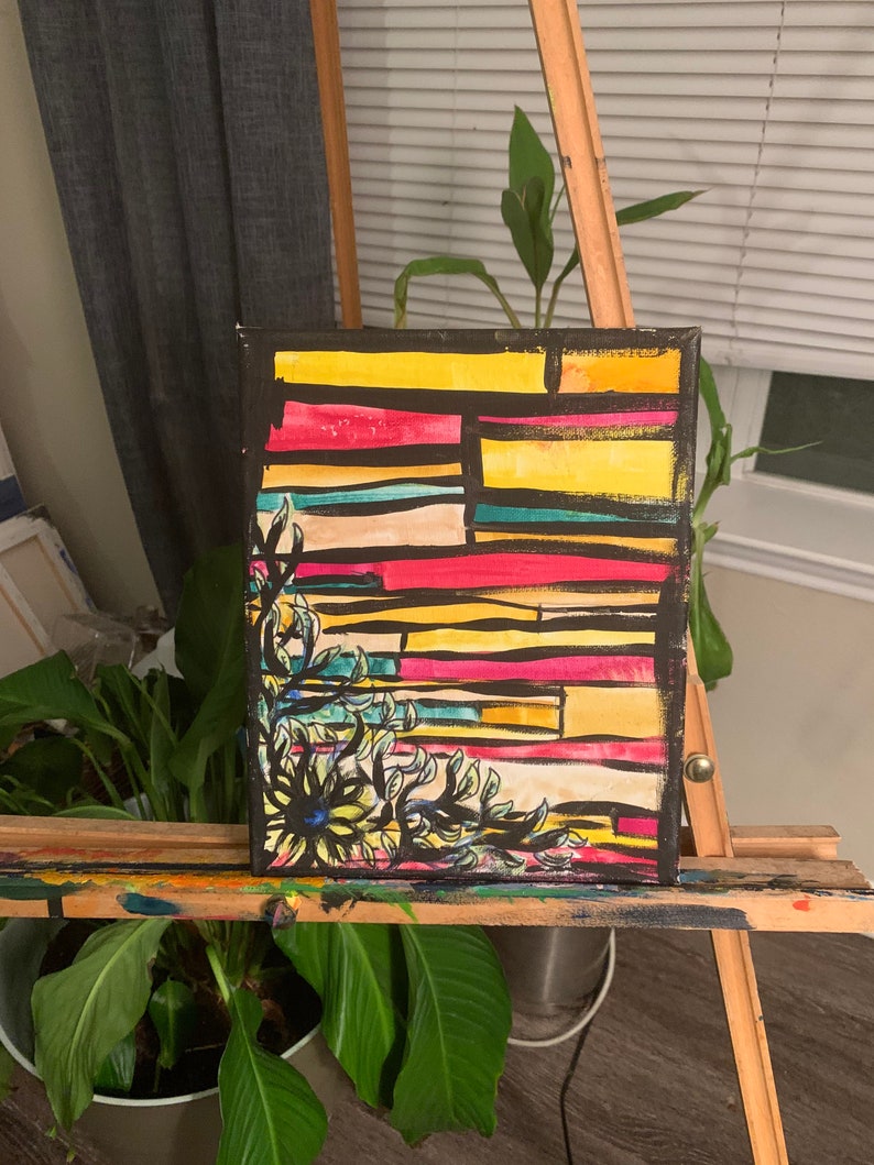 May include: Abstract art painting on canvas. The artwork features a colorful geometric design with yellow, red, and blue horizontal stripes. A black floral design is in the lower left corner. The painting is displayed on a wooden easel.