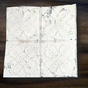May include: A white, distressed, tin ceiling tile with a floral pattern. The tile has a square shape and is made of metal.