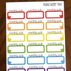 May include: A set of 18 colorful rectangular stickers with a dashed border and a small heart in the corner. Each sticker says "monthly pick" in black cursive writing. The stickers are in the colors red, orange, yellow, green, blue, purple, and pink.