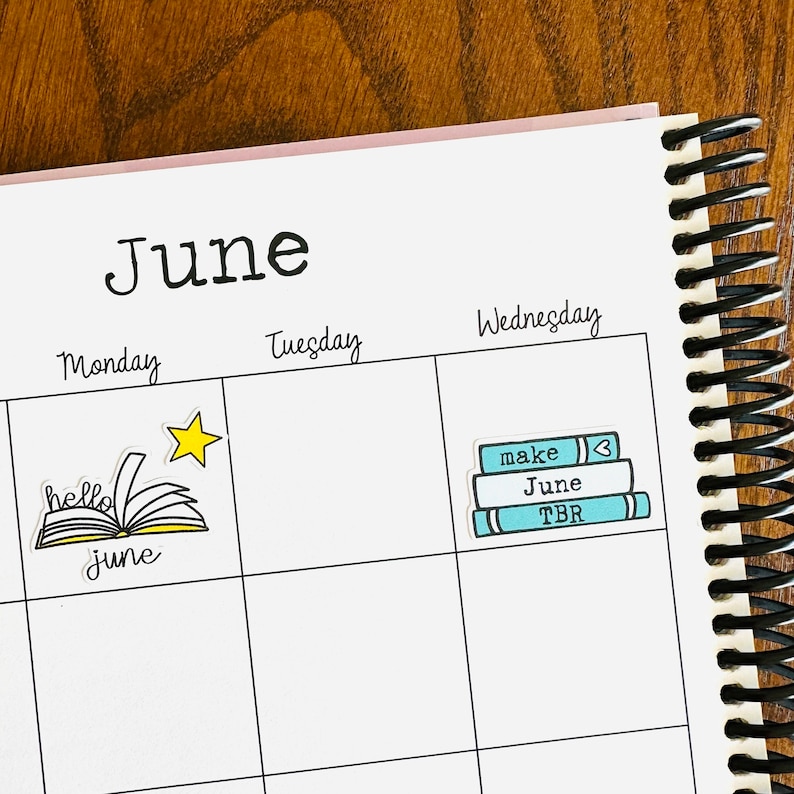May include: A June calendar page with a sticker of a stack of books with the words "make", "June", and "TBR" on them. The sticker is on the Wednesday box. The Monday box has a sticker of a book with the words "hello june" on it.