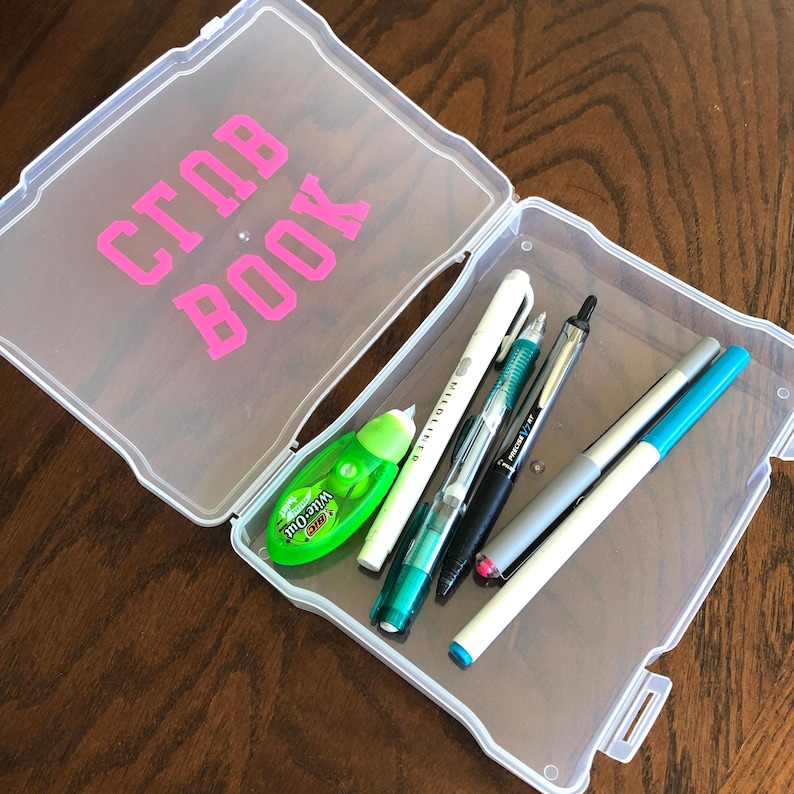 May include: A clear plastic storage box with the words "Club Book" written in pink letters on the lid. The box is open and contains a variety of pens, pencils, and a green tape dispenser.
