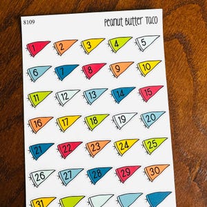May include: A white sticker sheet labeled Peanut Butter Taco 8109 featuring thirty-one colorful pennant-shaped stickers. Each pennant is numbered sequentially from 1 to 31 in black font, showcasing a variety of vibrant colors including red, orange, yellow, lime green, and shades of blue.