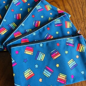 May include: A collection of blue zippered pouches with a repeating pattern of colorful stacked books and stars. The pouches have black zippers and white stitching. The books are in shades of red, orange, yellow, green, blue, and purple.
