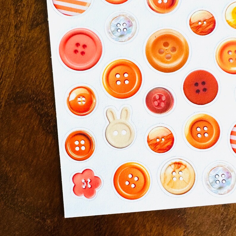 May include: A sheet of stickers featuring various orange and red button designs. The stickers include round buttons with four holes, a flower-shaped button, and a bunny-shaped button. The buttons vary in shade from light peach to deep red.