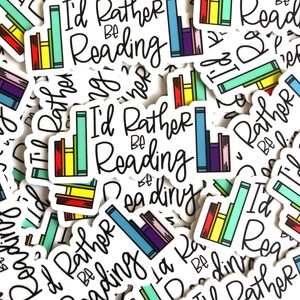 Op de afbeelding: Een close-up van verschillende stickers met de tekst "I'd Rather Be Reading" en een stapel kleurrijke boeken. De stickers zijn wit met zwarte tekst en kleurrijke boekillustraties.