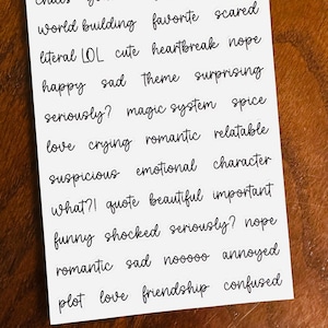 May include: A white sheet of paper with black handwritten words. The words are organized in rows and include words like "world building", "favorite", "scared", "literal LOL", "cute", "heartbreak", "nope", "happy", "sad", "theme", "surprising", "seriously?", "magic system", "spice", "love", "crying", "romantic", "relatable", "suspicious", "emotional", "character", "what?!", "quote", "beautiful", "important", "funny", "shocked", "seriously?", "nope", "romantic", "sad", "nooooo", "annoyed", "plot", "love", "friendship", "confused".