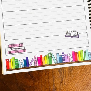 May include: A white lined notebook with a brown wooden cover. The notebook has a sticker of a stack of books with the words "make", "November", and "TBR" on it. There is also a sticker of an open book on the page.