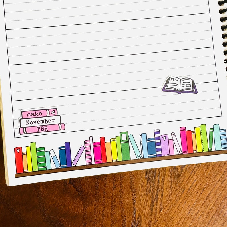 May include: A white lined notebook with a brown wooden cover. The notebook has a sticker of a stack of books with the words "make", "November", and "TBR" on it. There is also a sticker of an open book on the page.