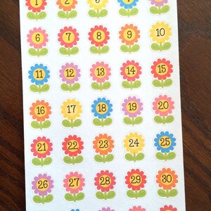 May include: A sheet of 31 colorful flower stickers with numbers 1 through 31. The flowers are yellow, orange, red, pink, purple, and blue. The stickers are on a white background. Peanut Butter Taco is written at the top of the sheet.