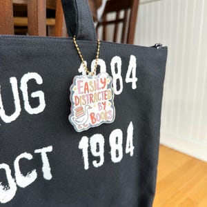 May include: Black tote bag with white text and a clear acrylic charm. The charm reads "Easily Distracted by Books" with a cartoon goose and books. The bag also has the numbers "1984" printed on it.