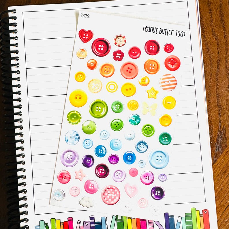 May include: A sheet of colorful buttons in various sizes and shapes, including hearts, stars, and flowers, arranged in a rainbow spectrum. The text "Peanut Butter Taco" is visible. The sheet is on a spiral-bound notebook with lined pages.