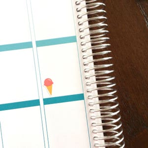 May include: A close-up of a white planner with a silver coil binding. A pink and orange ice cream cone sticker is on the page.
