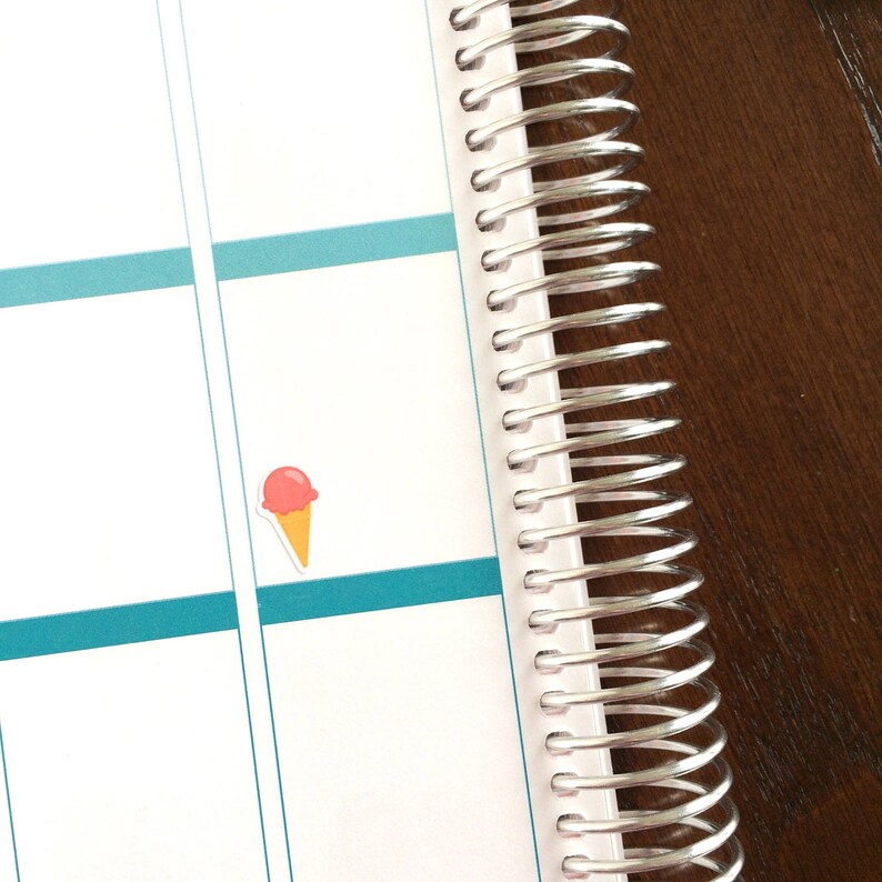 May include: A close-up of a white planner with a silver coil binding. A pink and orange ice cream cone sticker is on the page.