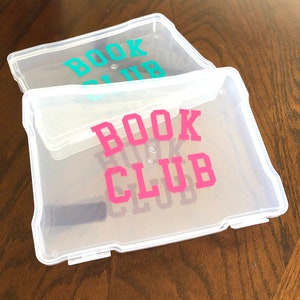 May include: Two clear plastic storage boxes with the words "BOOK CLUB" written on them. The top box has the words in teal, and the bottom box has the words in pink.