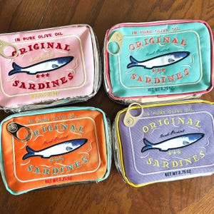May include: Four colorful, rectangular pouches resembling sardine cans. Each pouch features embroidered text reading "ORIGINAL SARDINES" and a fish illustration. The pouches are in pink, teal, orange, and purple, with a net weight of 3.75 oz.