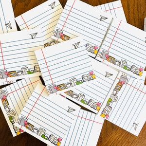 May include: A stack of lined paper notepads with colourful illustrations of books, coffee cups, and hearts. The notepads are ideal for taking notes, writing lists, or leaving messages.